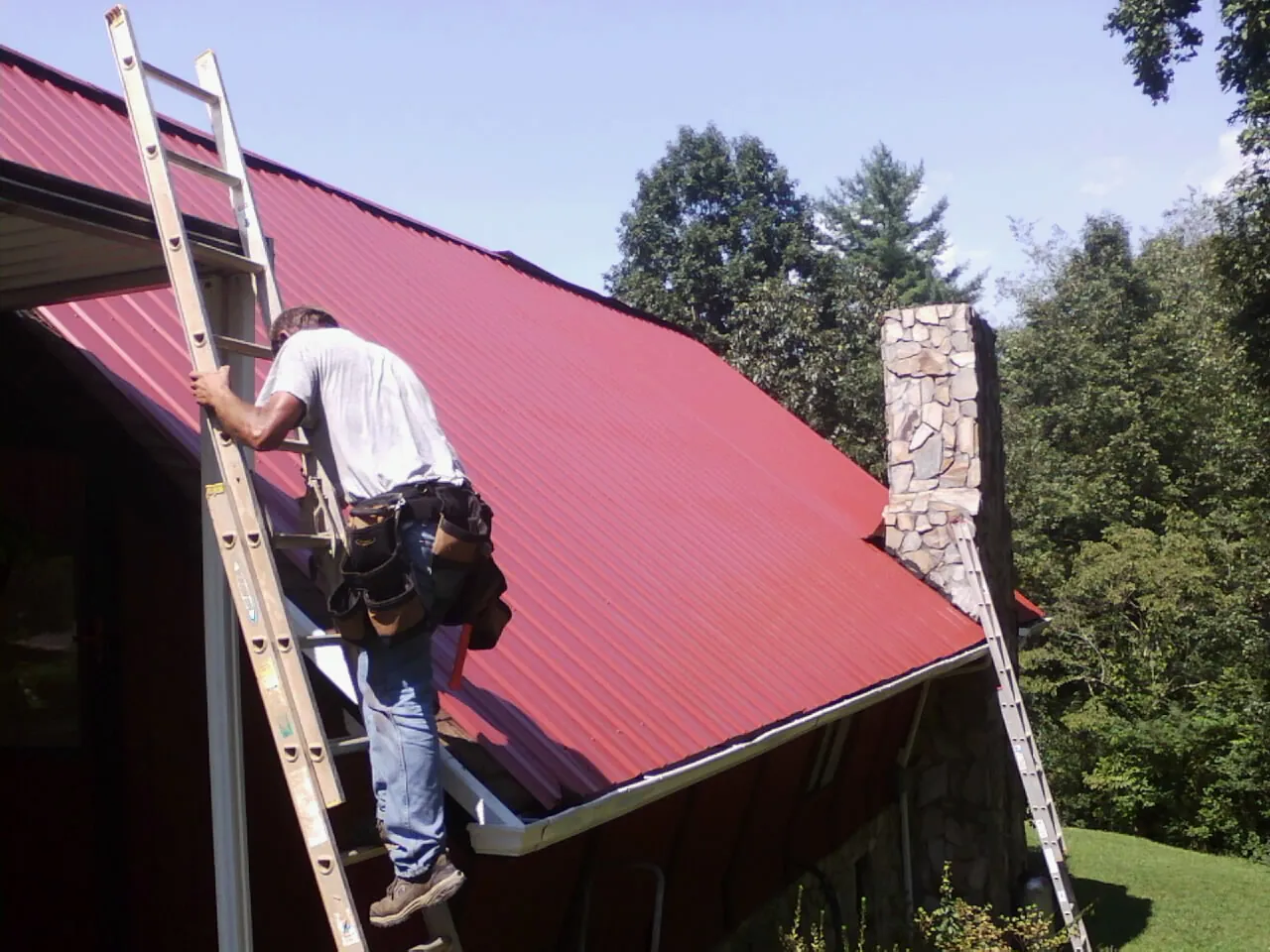 Roofer on a ladder beside a red metal roof and stone chimney — Soffit & Fascia Repair by Roofing Brothers in Searchlight